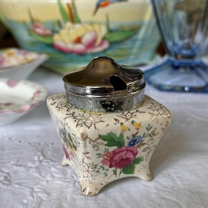 May include: A small, square, white ceramic salt or pepper shaker with a floral pattern and gold accents. The shaker has a silver lid with a small opening for pouring.