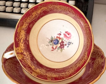 May include: A vintage teacup and saucer set. The cup and saucer have a burgundy rim and gold floral detailing. The center of the cup features a floral bouquet. The set is placed on a white surface.