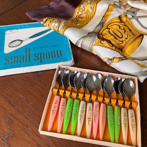 Könnte beinhalten: Ein Set aus zwölf kleinen Edelstahllöffeln in einer rechteckigen Schachtel. Jeder Löffel hat eine metallische Laffe und einen schlanken Griff in Pastellrosa, Limettengrün oder Creme, verziert mit einem goldenen Blumenmuster. Der Deckel zeigt den Text small spoon.