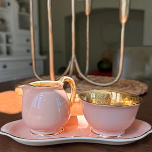 Puede incluir: Un juego de crema y azucarero de porcelana rosa con ribetes dorados, sobre una bandeja a juego. El juego se exhibe sobre una superficie de madera oscura, con un candelabro dorado y velas de colores en el fondo.