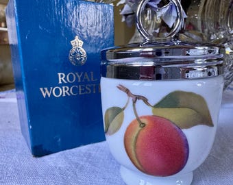 Könnte beinhalten: Eine weiße Keramik-Eierbecher mit einem silbernen Deckel und Griff. Der Eierbecher hat ein Bild einer roten Pflaume mit grünen Blättern. Der Eierbecher befindet sich in seiner Originalverpackung, die blau ist und die Worte "Royal Worcester" aufgedruckt hat.