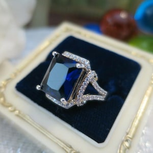 May include: A silver ring featuring a large, rectangular, deep blue gemstone. The ring is adorned with smaller, clear stones around the main stone and on the band, displayed in a velvet-lined box.