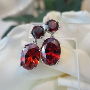 May include: A pair of dangle earrings with red oval-shaped gemstones and smaller round red gemstones at the top.