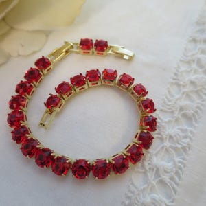May include: A gold bracelet featuring a series of bright red gemstones. The bracelet is displayed on a white surface, and the gemstones are oval-shaped. The clasp is visible. The bracelet is new.