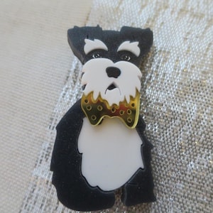 May include: A Schnauzer dog-shaped pin in black, white, and gold. The pin has a black body, a white chest and beard, and a gold bow tie. The dog is depicted in a sitting position.