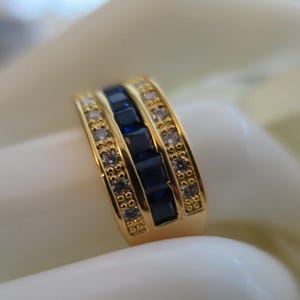 May include: A gold band ring featuring a central row of square-cut deep blue stones flanked by two outer rows of small, round, clear white stones. The metal has a polished finish, and the design incorporates a channel setting for the blue stones.