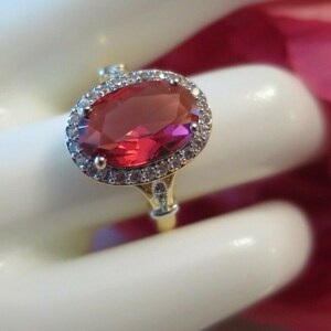 May include: A gold ring with a large, oval-shaped, red gemstone, encircled by a halo of small, clear stones. The ring's band is also adorned with smaller stones. The ring is shown against a soft, white background, highlighting its sparkle.