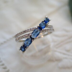 May include: A silver ring with a double band design. One band is encrusted with small, clear stones, while the other features a row of five oval, deep blue gemstones. The ring is set against a soft, white background.