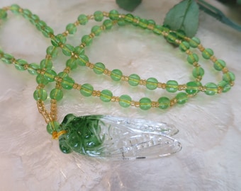 May include: A green and clear glass pendant necklace with a detailed cicada design. The necklace is made with green and gold beads.