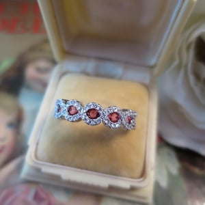 May include: A silver-toned ring featuring a row of five round-cut red gemstones, each surrounded by a halo of small clear stones, set within a cream-coloured velvet-lined jewellery box.