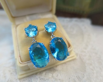 May include: A pair of silver-toned drop earrings with large, oval, light blue gemstones. Each earring features a smaller, round gemstone at the top. The earrings are displayed in a vintage, cream-colored jewelry box.