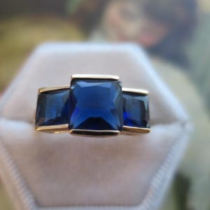 May include: A gold ring featuring three deep blue rectangular-cut stones arranged in a row. The centre stone is larger than the two flanking side stones, all set within a smooth gold band that holds the gems securely in a horizontal alignment.