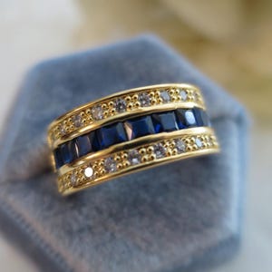 May include: A gold band ring featuring a central row of square-cut deep blue stones flanked by two rows of small, clear, round-cut stones. The metal has a polished finish, and the design incorporates a repeating beaded texture along the edges.