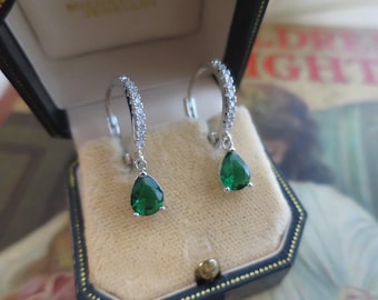 May include: A pair of silver earrings with a teardrop-shaped, emerald-green gemstone pendant. The earrings are displayed in a black jewelry box with a beige interior. The hoops are encrusted with small, clear gemstones.