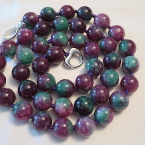 May include: A necklace with large, round beads in shades of deep purple and teal green. The beads are strung on a purple cord and secured with a silver-tone clasp. A silver heart-shaped charm is also included.