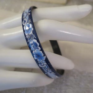 May include: A slim bangle bracelet featuring a dark navy blue base with a repeating pattern of light blue and white flowers and delicate green leaves. The floral design wraps around the exterior surface of the smooth, glossy circular band.