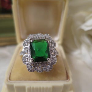 May include: A silver-toned ring featuring a large, rectangular, emerald-green centre stone surrounded by a double halo of small, clear, round-cut stones. The setting has a scalloped, floral-inspired edge, creating a decorative, layered appearance.