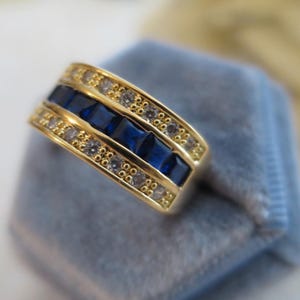 May include: A gold-toned band ring featuring a central row of five square-cut deep blue stones flanked by two outer rows of small, clear round stones. The design is symmetrical, with the blue stones set in a channel between the clear stone rows.