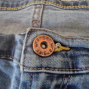 K&ouml;nnte beinhalten: Nahaufnahme einer blauen Jeans mit einem kupferfarbenen Knopf. Der Knopf ist mit dem Wort "Wrangler" gepr&auml;gt. Die Jeans hat ein "RELAXED"-Etikett und gelbe N&auml;hte.