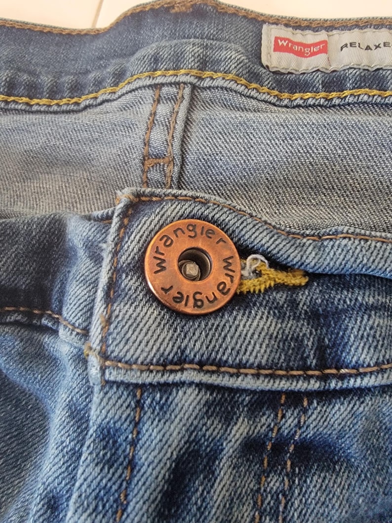 K&ouml;nnte beinhalten: Nahaufnahme einer blauen Jeans mit einem kupferfarbenen Knopf. Der Knopf ist mit dem Wort "Wrangler" gepr&auml;gt. Die Jeans hat ein "RELAXED"-Etikett und gelbe N&auml;hte.