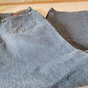 May include: Light blue denim jeans with a classic five-pocket design. The jeans have a faded wash and a straight leg cut. The back pocket features a small, rectangular patch. The jeans are folded on a white surface.