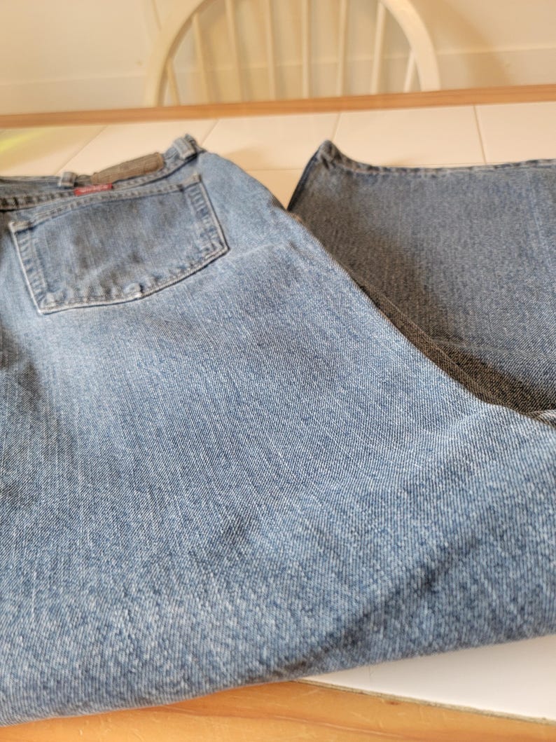 May include: Light blue denim jeans with a classic five-pocket design. The jeans have a faded wash and a straight leg cut. The back pocket features a small, rectangular patch. The jeans are folded on a white surface.