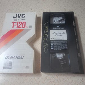 May include: A white JVC T-120 Dynarec VHS video cassette sleeve with red and orange diagonal stripes next to a black Scotch VHS tape labeled A Clockwork Orange by Warner Home Video, featuring handwritten text on the plastic casing.