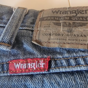 May include: Close-up of blue denim jeans with the Wrangler brand label. The label is brown with the words "Wrangler PREMIUM QUALITY" and an eagle graphic. A red tag with the Wrangler logo is sewn on the jeans.