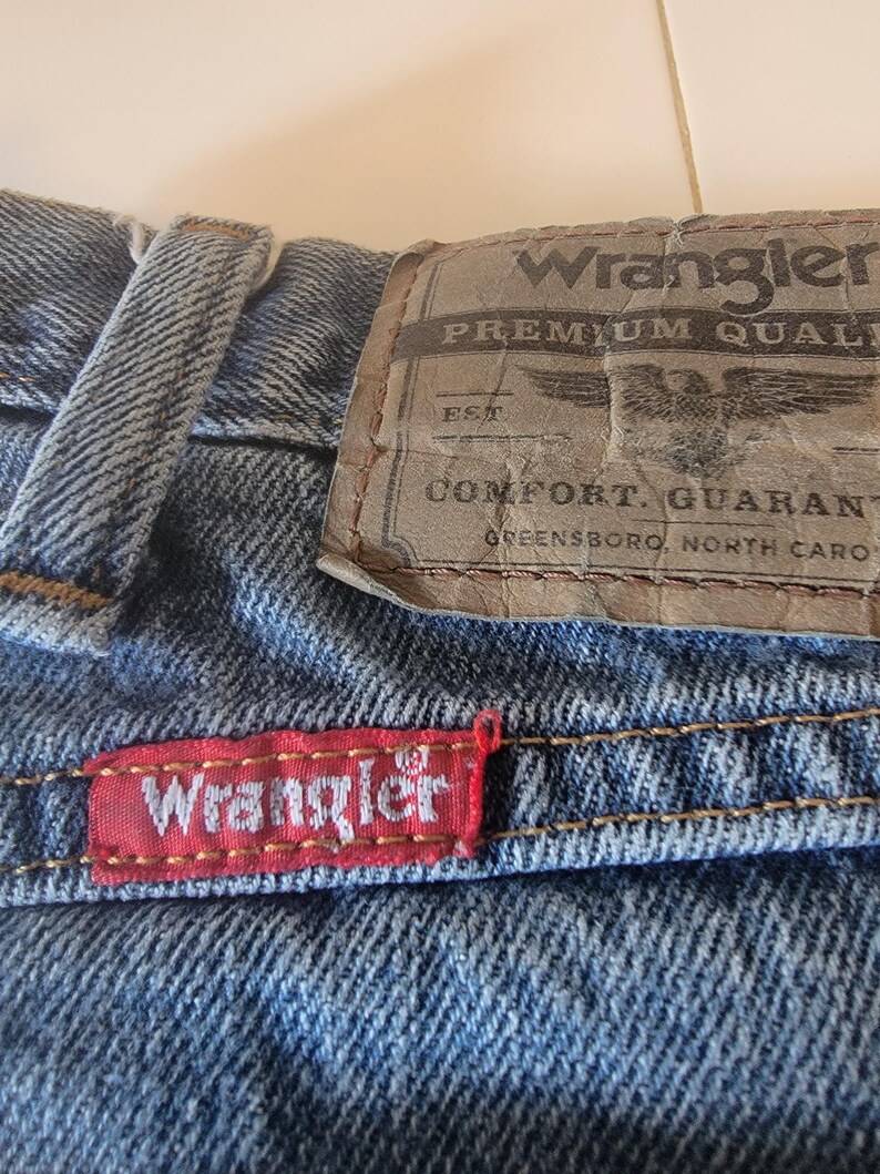May include: Close-up of blue denim jeans with the Wrangler brand label. The label is brown with the words "Wrangler PREMIUM QUALITY" and an eagle graphic. A red tag with the Wrangler logo is sewn on the jeans.
