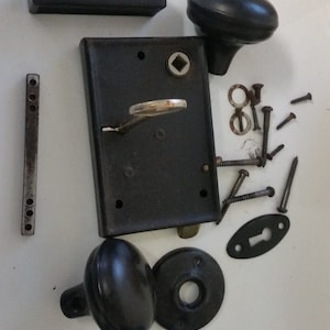 May include: A vintage door hardware set in black with two round knobs, a rectangular plate with a keyhole, a strike plate, and a set of screws.
