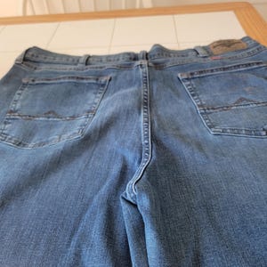 K&ouml;nnte beinhalten: Blaue Jeans aus Denim mit Ges&auml;&szlig;taschen und einem Lederaufn&auml;her. Die Jeans bestehen aus einem Denim-Stoff mit mittlerer Waschung. Die Ges&auml;&szlig;taschen haben ein dekoratives Nahtdesign. Die Jeans liegen flach auf einer Oberfl&auml;che.