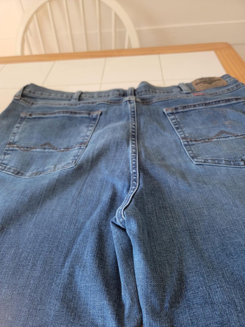 K&ouml;nnte beinhalten: Blaue Jeans aus Denim mit Ges&auml;&szlig;taschen und einem Lederaufn&auml;her. Die Jeans bestehen aus einem Denim-Stoff mit mittlerer Waschung. Die Ges&auml;&szlig;taschen haben ein dekoratives Nahtdesign. Die Jeans liegen flach auf einer Oberfl&auml;che.