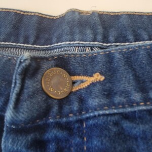 May include: Close-up of a pair of blue denim jeans. The jeans feature a button closure with the brand name "Wrangler" embossed on the button. Visible stitching and a buttonhole are also present.