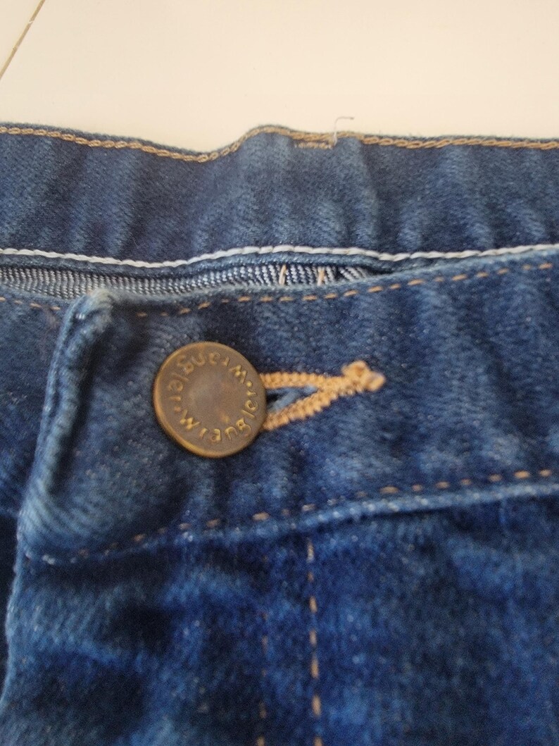 May include: Close-up of a pair of blue denim jeans. The jeans feature a button closure with the brand name "Wrangler" embossed on the button. Visible stitching and a buttonhole are also present.
