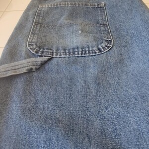 May include: Close-up of blue denim jeans. The jeans have a classic five-pocket design, with a large back pocket visible. The denim fabric has a textured appearance, and the stitching is visible along the seams and pocket edges. The jeans are folded.