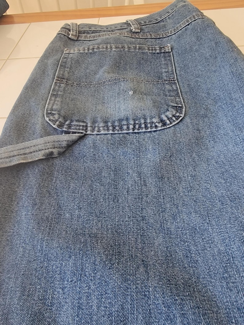 May include: Close-up of blue denim jeans. The jeans have a classic five-pocket design, with a large back pocket visible. The denim fabric has a textured appearance, and the stitching is visible along the seams and pocket edges. The jeans are folded.