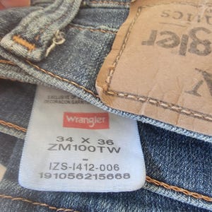 May include: Close-up of blue denim jeans with a Wrangler label. The label displays the size 34 x 36, along with other product information. The jeans have a classic five-pocket design and a leather patch.