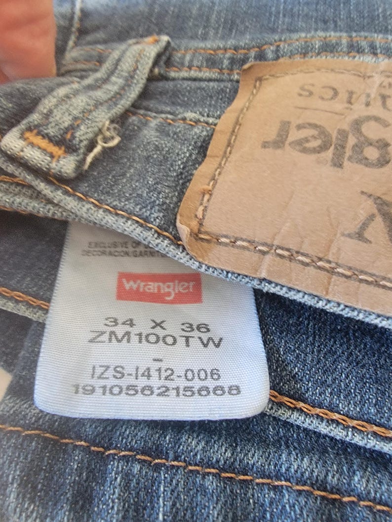 May include: Close-up of blue denim jeans with a Wrangler label. The label displays the size 34 x 36, along with other product information. The jeans have a classic five-pocket design and a leather patch.