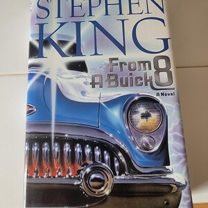 May include: A blue and silver car with a chrome bumper and a red and orange glow coming from the grill. The book cover reads "Stephen King From A Buick 8 A Novel".