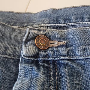 May include: Close-up of blue denim jeans with a button and buttonhole. The button is bronze-colored and has the word "Wrangler" around the edge. The denim has a faded appearance.