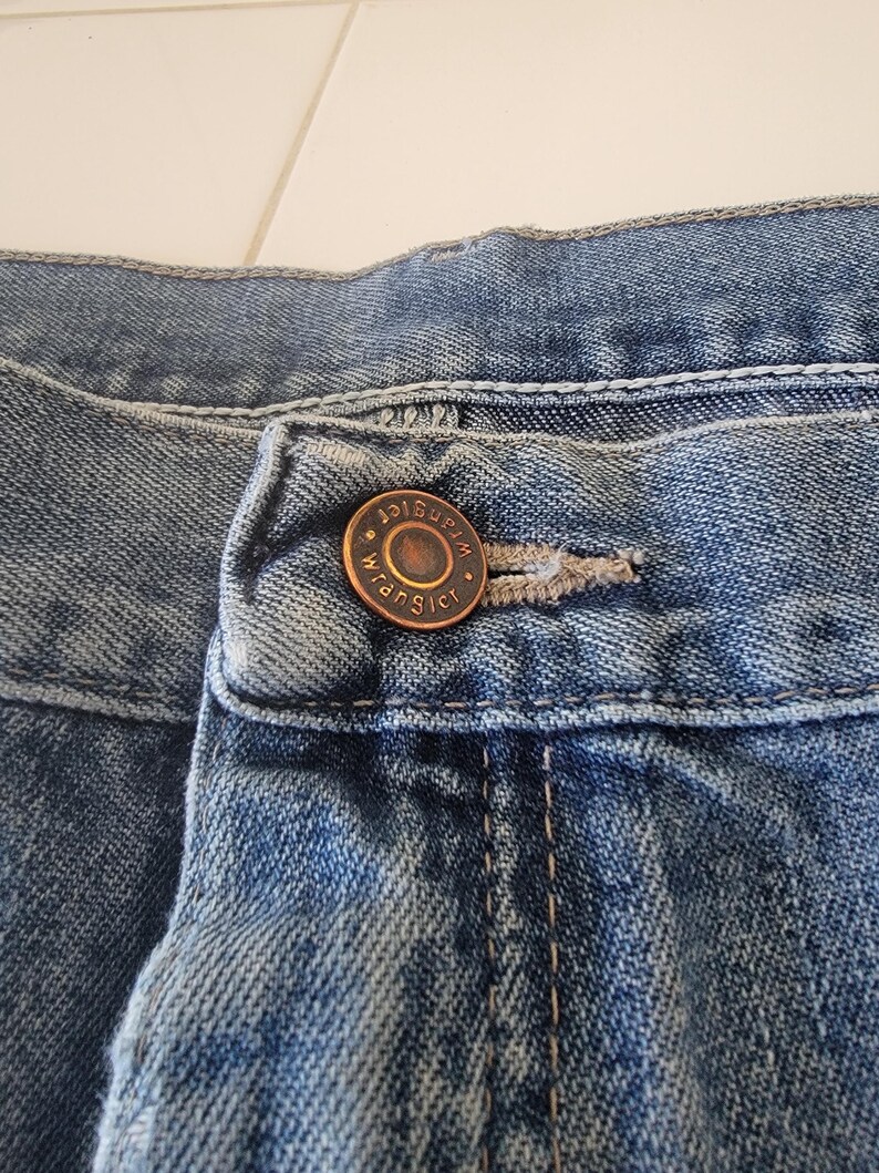 May include: Close-up of blue denim jeans with a button and buttonhole. The button is bronze-colored and has the word "Wrangler" around the edge. The denim has a faded appearance.