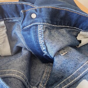 May include: Close-up of blue denim jeans, showing the waistband, button, and stitching details. The jeans have a classic five-pocket design and a faded wash. The stitching is white, and the button is silver.