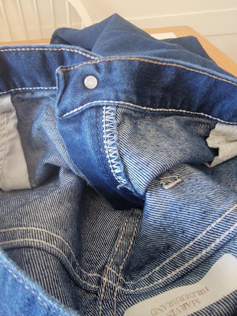 May include: Close-up of blue denim jeans, showing the waistband, button, and stitching details. The jeans have a classic five-pocket design and a faded wash. The stitching is white, and the button is silver.