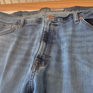 K&ouml;nnte beinhalten: Nahaufnahme einer blauen Jeans. Die Jeans hat einen Knopfverschluss, G&uuml;rtelschlaufen und Taschen. Der Denim hat ein verwaschenes Aussehen mit helleren Bereichen an den Oberschenkeln und Knien. Die Jeans ist &uuml;ber eine Holzoberfl&auml;che gefaltet.