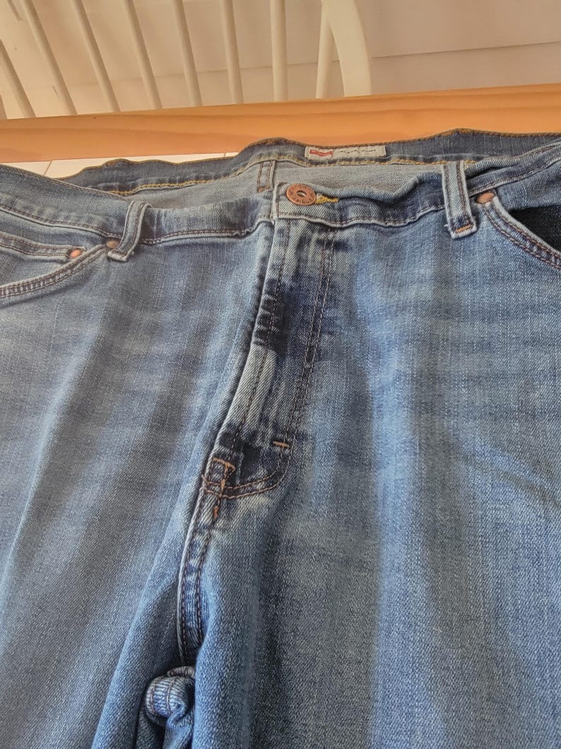 K&ouml;nnte beinhalten: Nahaufnahme einer blauen Jeans. Die Jeans hat einen Knopfverschluss, G&uuml;rtelschlaufen und Taschen. Der Denim hat ein verwaschenes Aussehen mit helleren Bereichen an den Oberschenkeln und Knien. Die Jeans ist &uuml;ber eine Holzoberfl&auml;che gefaltet.