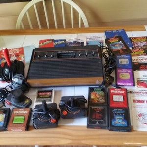 May include: A vintage Atari 2600 video game console with multiple game cartridges, controllers, and instruction manuals. The console is brown and black with a woodgrain finish. The cartridges are black with colorful labels. The controllers are black with red buttons. The manuals are printed on white paper with colorful graphics.