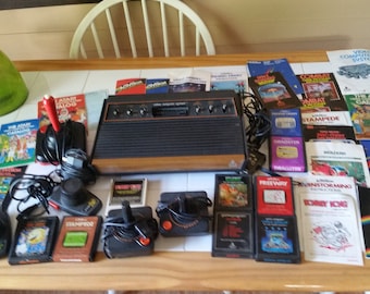 May include: A vintage Atari 2600 video game console with multiple game cartridges, controllers, and instruction manuals. The console is brown and black with a woodgrain finish. The cartridges are black with colorful labels. The controllers are black with red buttons. The manuals are printed on white paper with colorful graphics.
