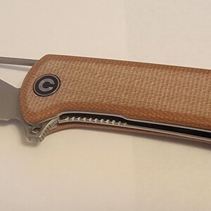 May include: A folding pocket knife with a tan handle and a silver blade. The blade has a curved shape with a cutout near the handle. The handle has a textured surface and a black and silver accent.