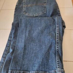 May include: Blue denim jeans with a classic five-pocket design. The jeans are folded, showcasing the back pocket and the texture of the denim fabric. The jeans have a button and zipper closure.
