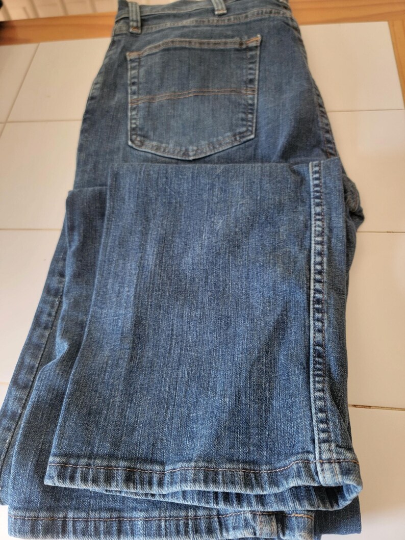 May include: Blue denim jeans with a classic five-pocket design. The jeans are folded, showcasing the back pocket and the texture of the denim fabric. The jeans have a button and zipper closure.